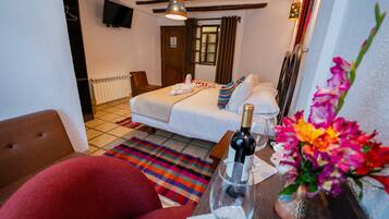 Habitación de hotel con una cama, un sillón rojo, una mesa de madera con una botella y un vaso, y una alfombra colorida.