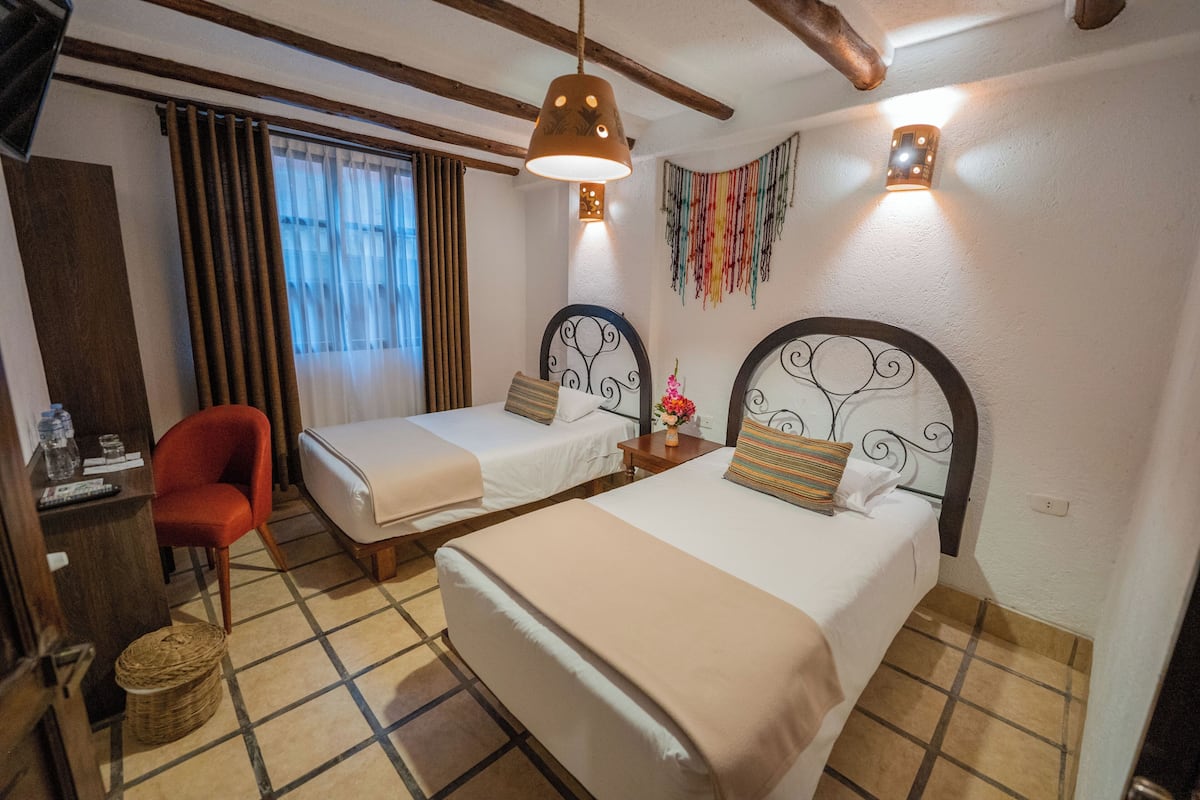 A hotel room with two single beds, a desk with a chair, a red armchair, and a decorative wall hanging.