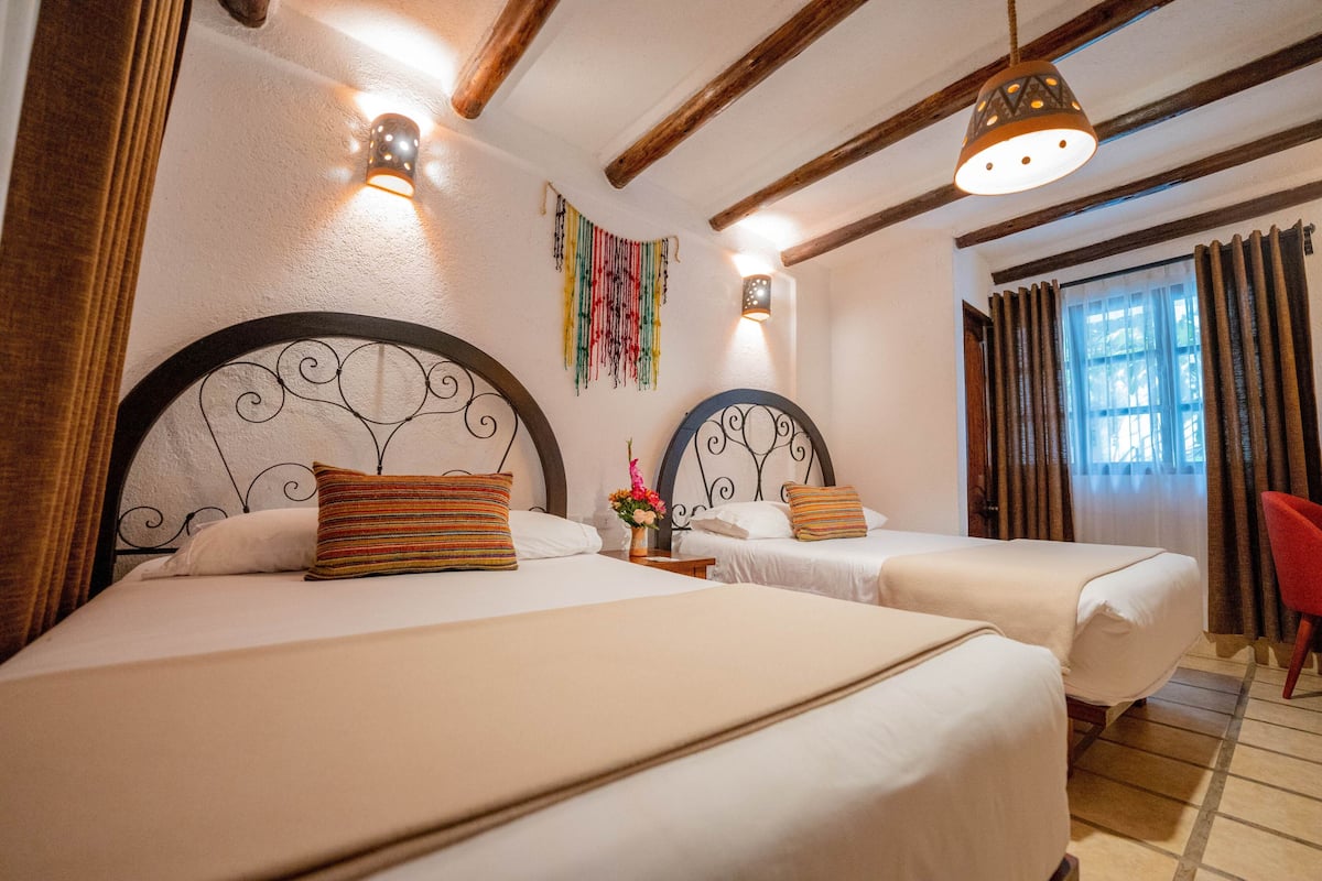 A hotel room with two beds, wooden beams on the ceiling, a red chair, and a window with curtains.