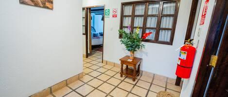 Un pasillo con piso de baldosas, una mesa de madera con una planta en maceta, un extintor y una puerta con un letrero rojo.