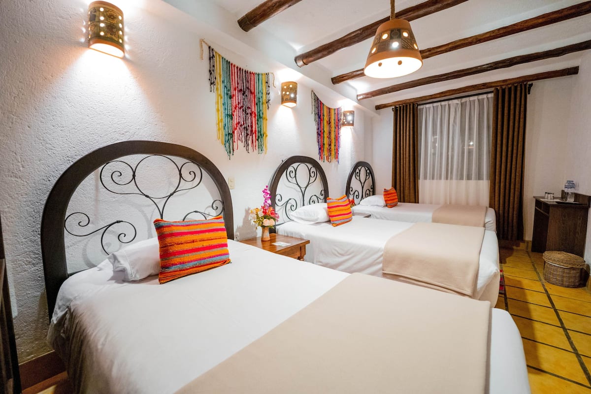 A hotel room with three beds, each with a unique headboard design, white bedding, and colorful pillows. There are wall-mounted lights and a window with curtains.