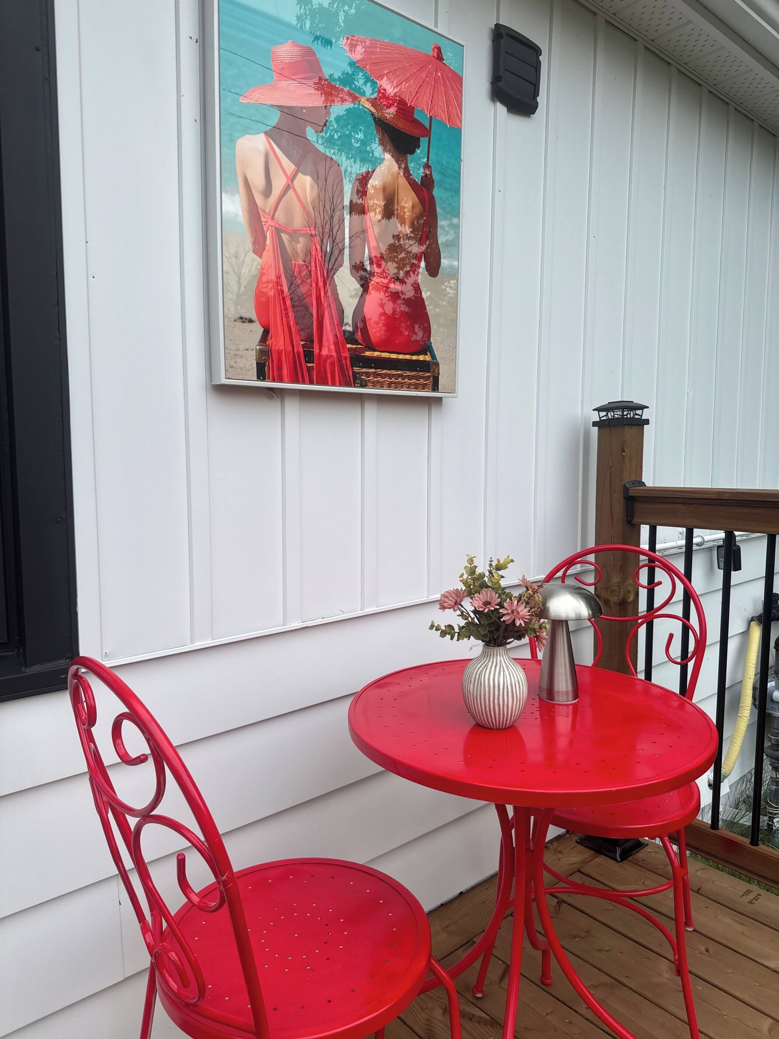A red metal table and chairs set on a wooden deck, with a framed picture on the wall.