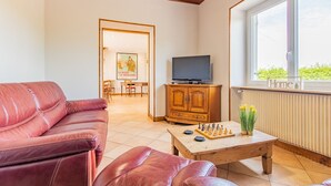 Ein Wohnzimmer mit einem roten Ledersofa, einem Couchtisch aus Holz mit Schachfiguren, einem Fernseher auf einer Holzkommode und einem Fenster mit einem Schild mit der Aufschrift 'Home'.