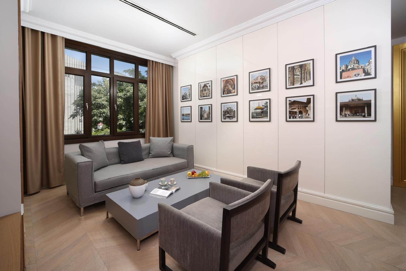 A modern living room with a sofa, coffee table, and chairs, a large window with curtains, and a wall with framed pictures.