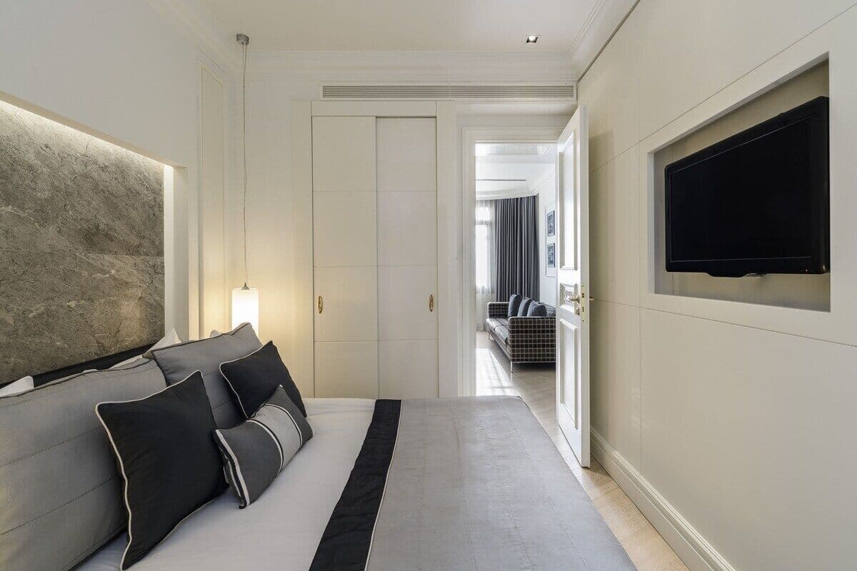 A modern bedroom with a large bed, a flat-screen TV mounted on the wall, and a view into the living area.