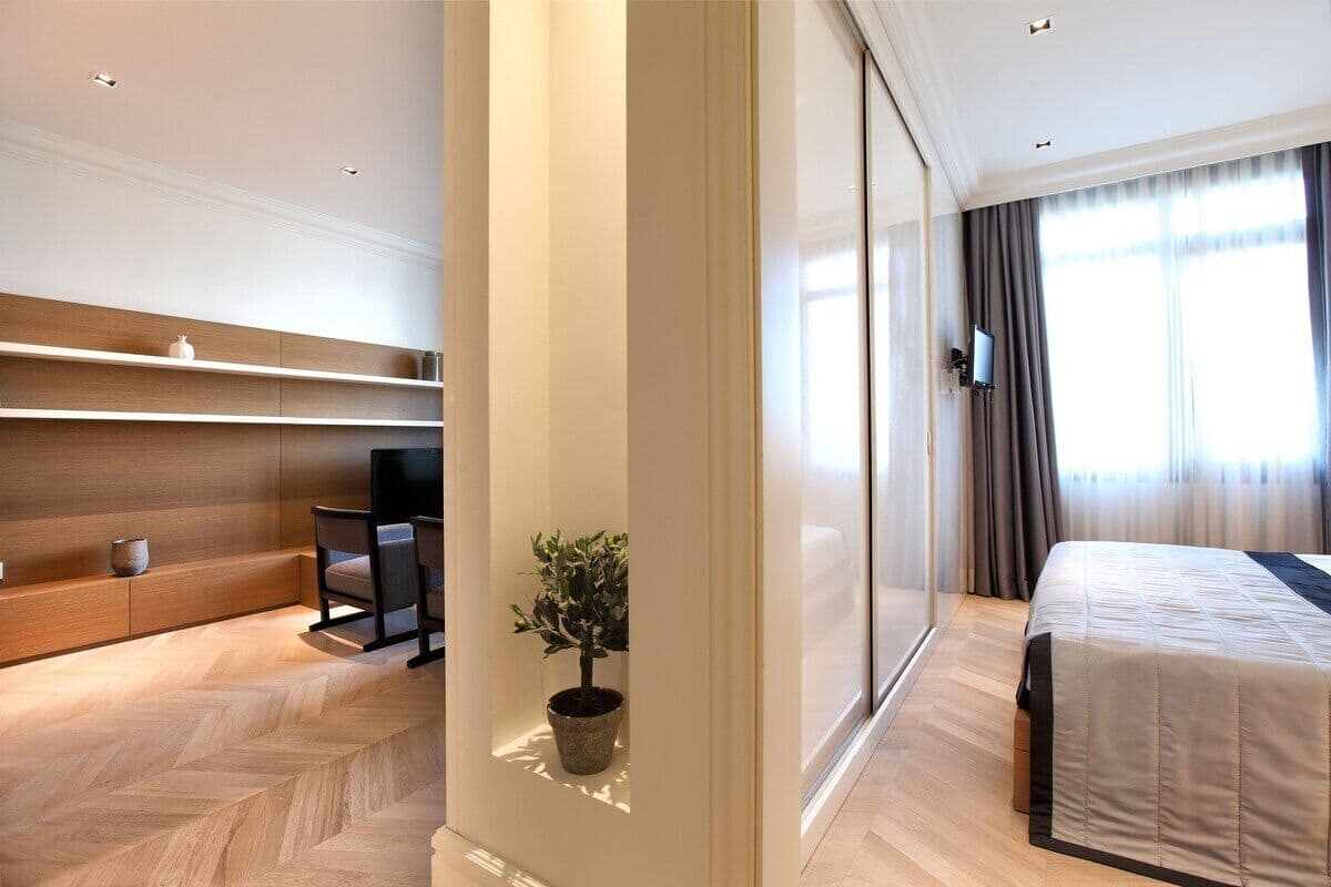 A modern hotel room with a bed, a seating area with a chair, a small potted plant, and a television mounted on the wall.