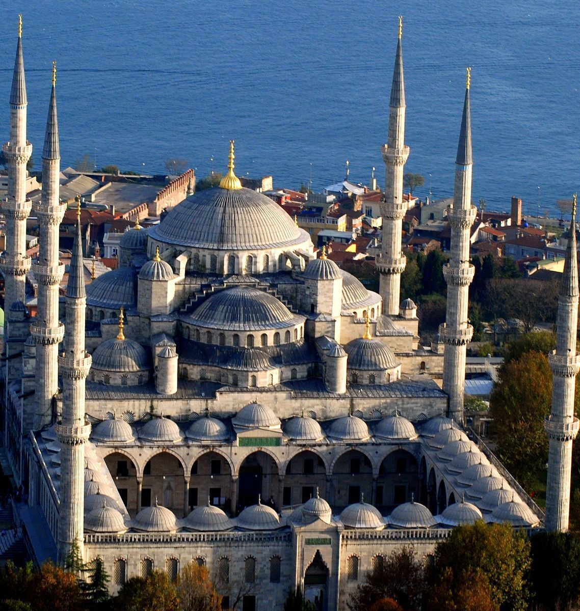 A large mosque with multiple domes and minarets, surrounded by a cityscape and a body of water.