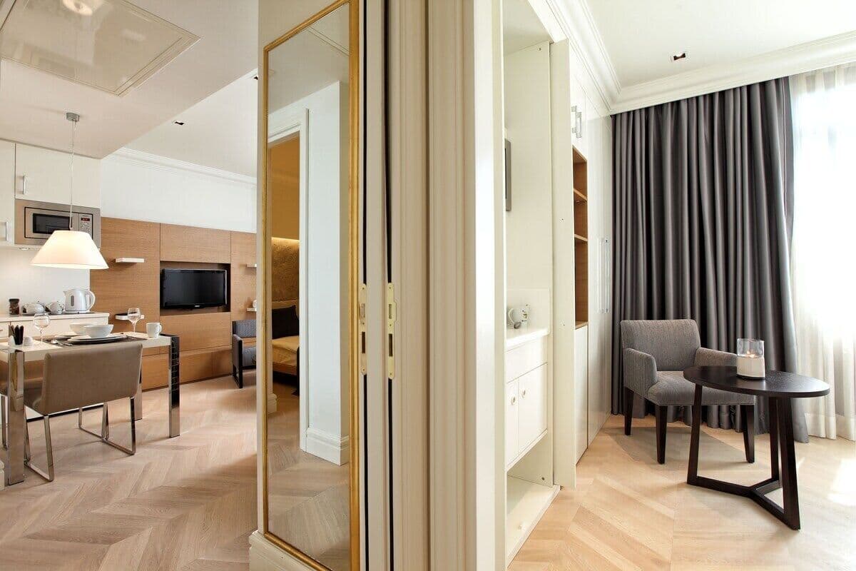 A modern hotel room with a dining area, a TV, a sofa, and a large window with curtains.