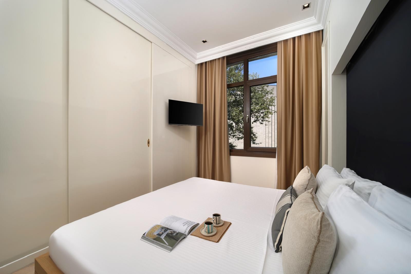 Habitación de hotel con cama, televisor de pantalla plana, ventana con cortinas y una revista sobre la cama.