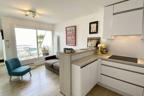 A modern kitchen with a built-in island, a dining area with a sofa, and a view through a large sliding glass door. - Appartement Avec Terrasse , Surplombant la Baie de Somme (Le Crotoy)