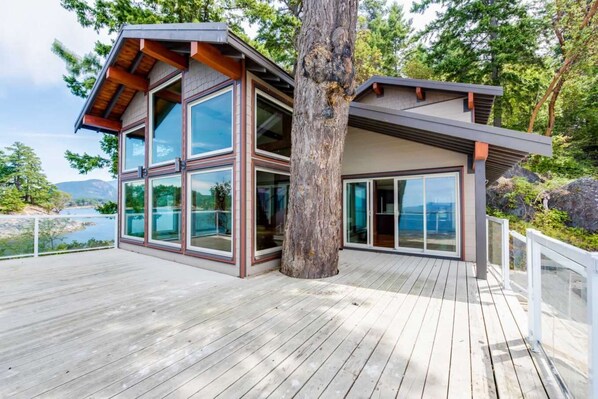 A modern house with large glass windows, a wooden deck, and a view of a lake. - Exclusive Private Island - Pacific Ocean Paradise (Madeira Park)