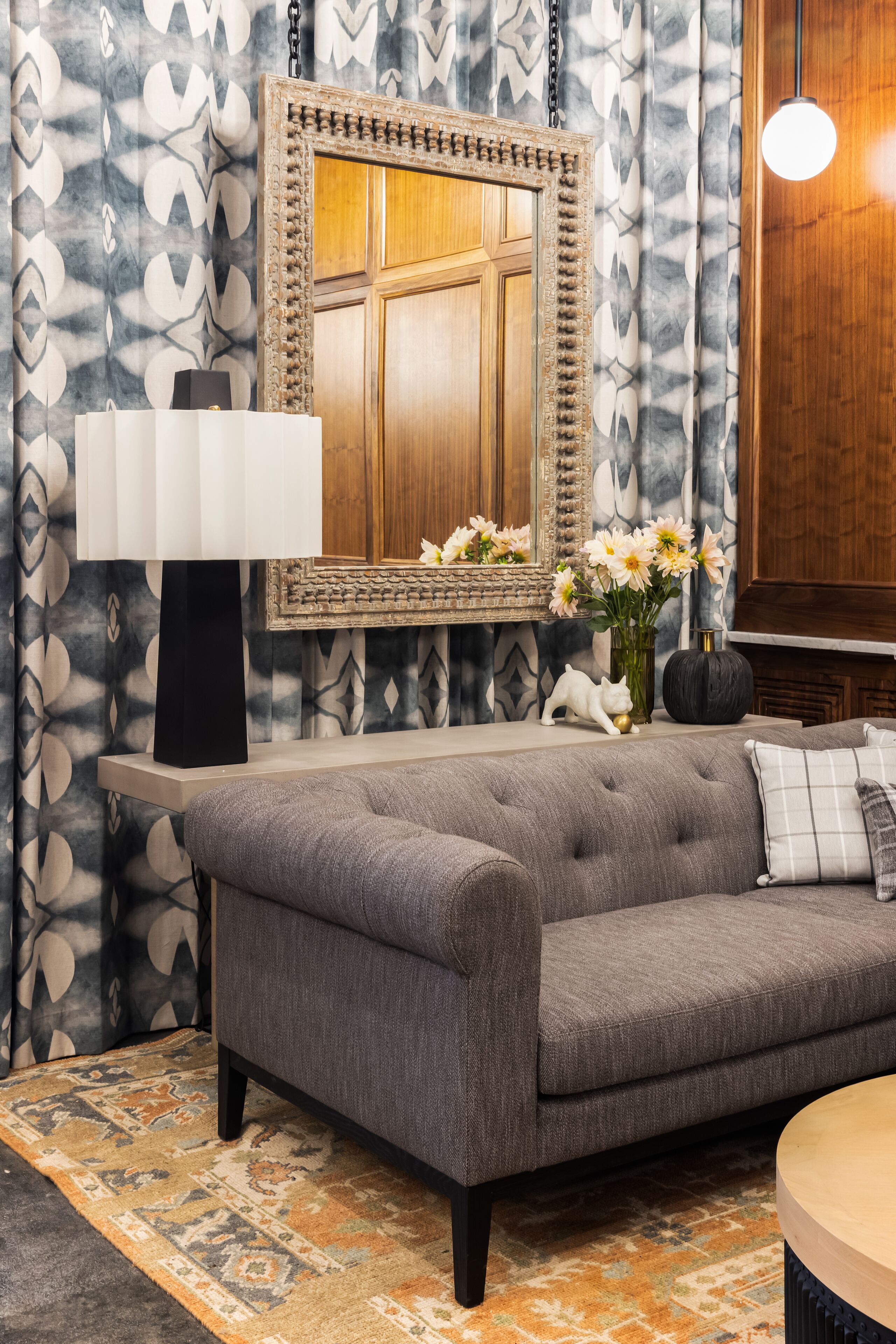 A modern living room with a grey sofa, a large mirror, a black lamp, and a vase with flowers.