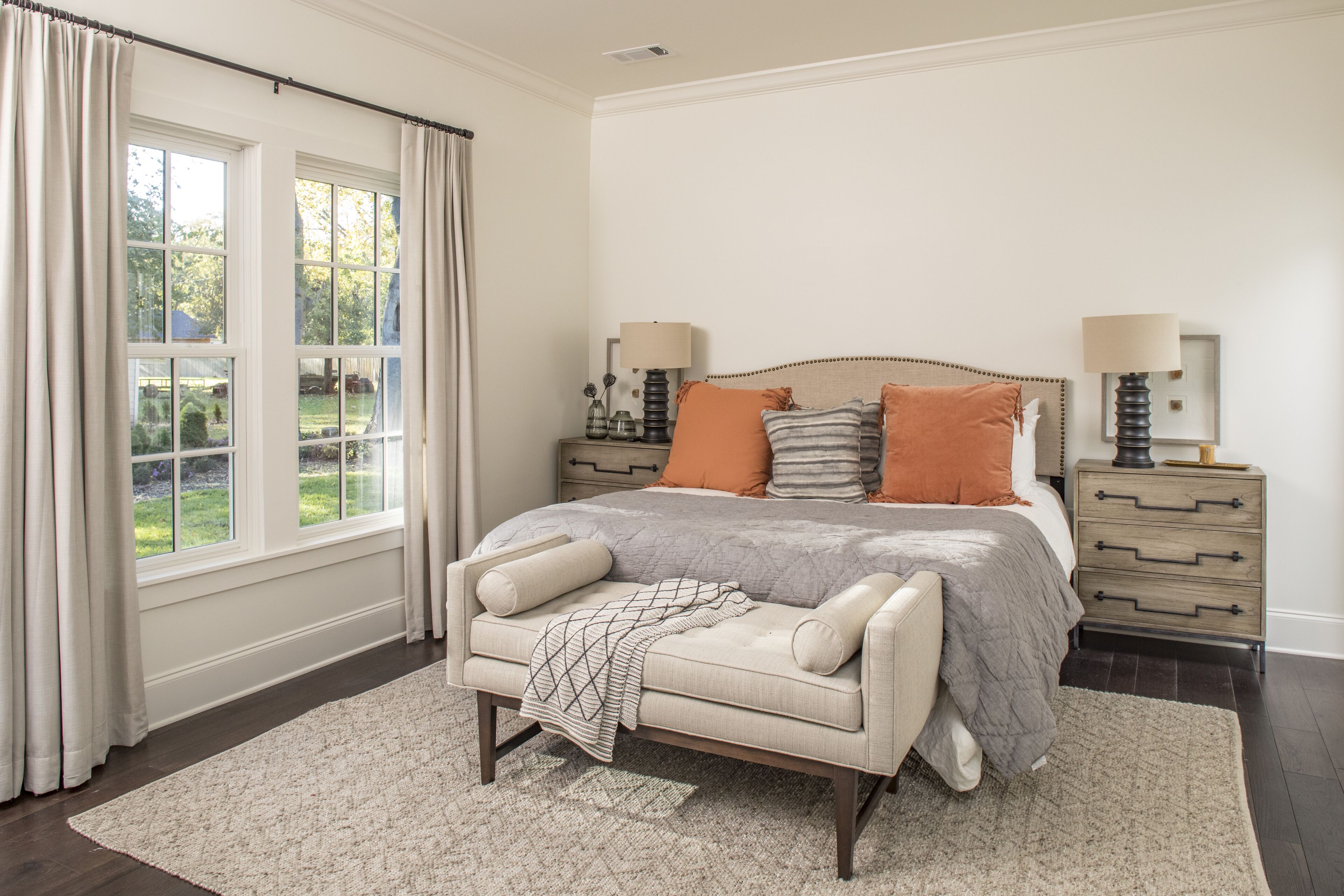 A bedroom with a large bed, two bedside tables with lamps, a bench, and a view of the outdoors through large windows.