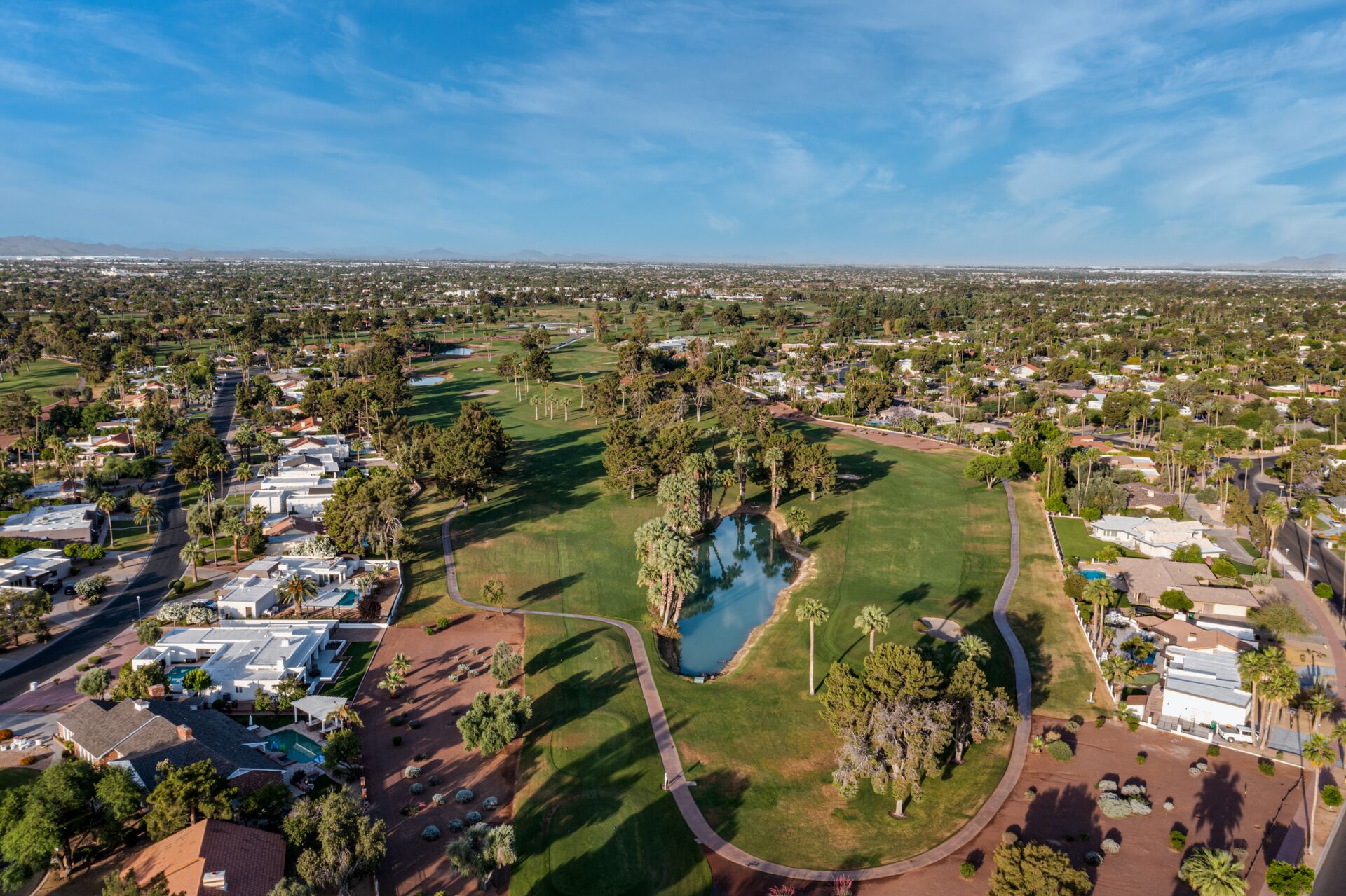 An aerial view of a golf course with a pond, surrounded by residential areas and a clear sky.