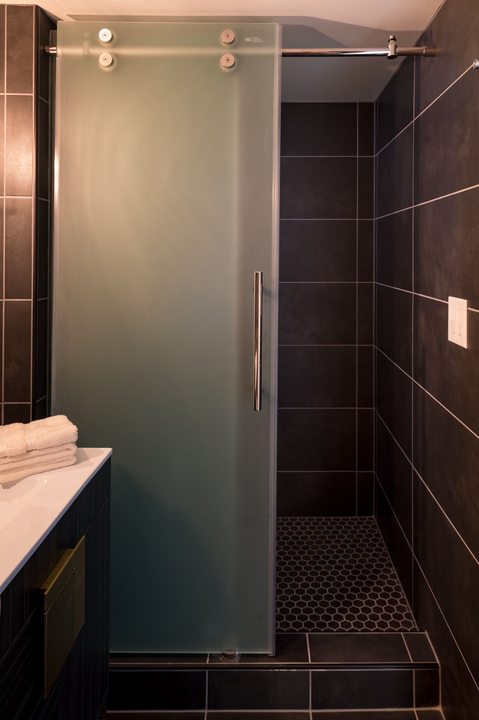Een moderne badkamer met een douchecabine van glas, donkere betegelde wanden en een wit aanrechtblad met handdoeken.