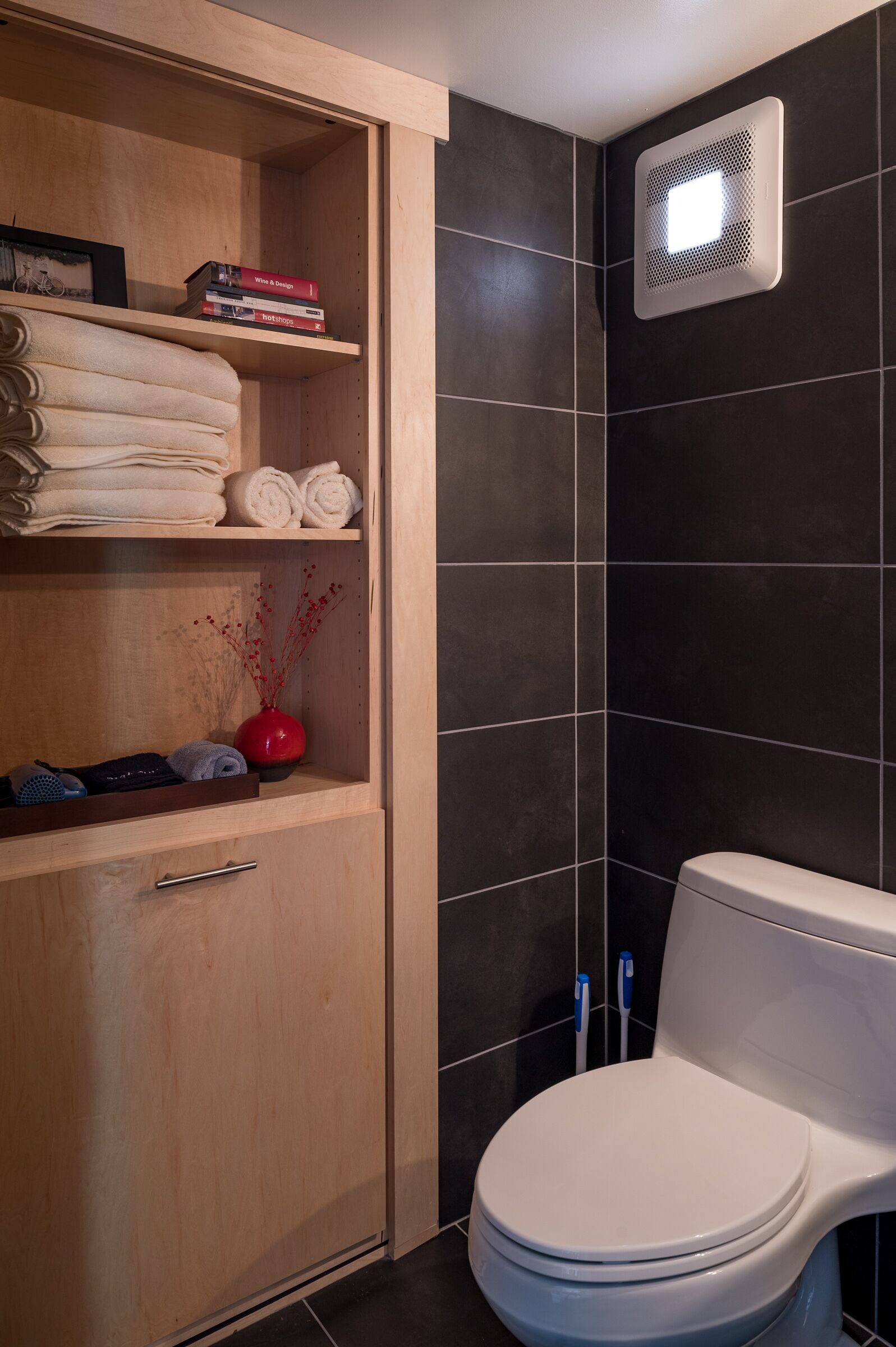 A bathroom with a white toilet, a wooden cabinet with towels, and a red vase.