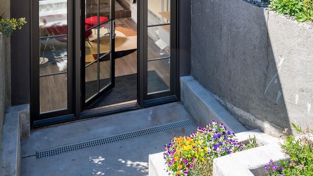 A modern building with a brick facade, large sliding glass doors leading to an outdoor seating area with red chairs, and a concrete planter with colorful flowers. - Queen Anne Design Studio, 7days max. (Seattle)