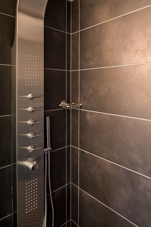A modern shower with multiple controls and a handheld showerhead, set against a dark tiled wall.