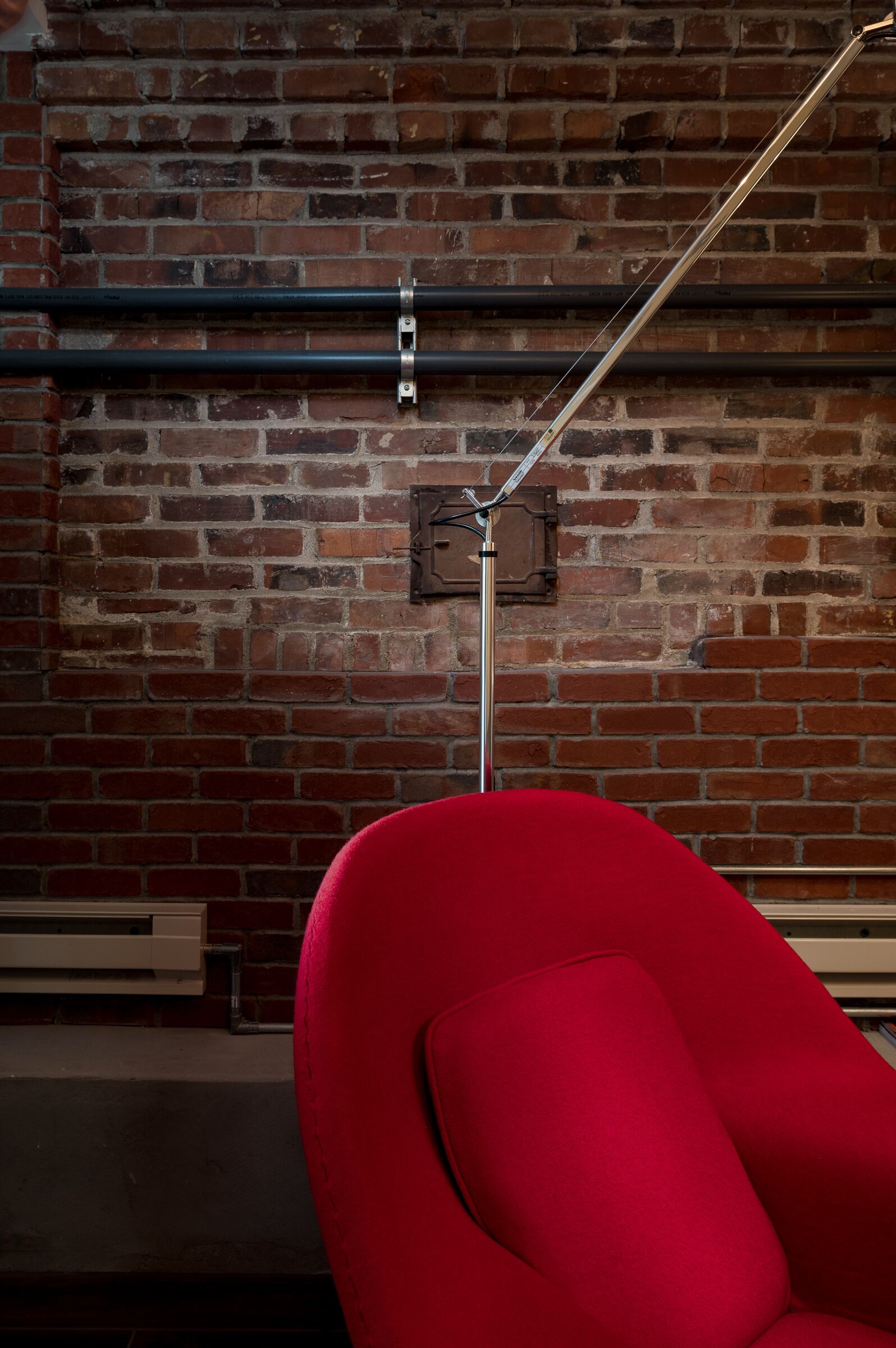 Une chaise rouge est placée devant un mur de briques, avec des tuyaux apparents et un luminaire en métal.