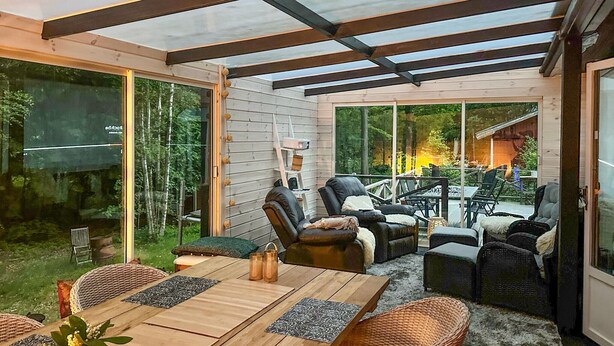 A covered patio with wooden furniture, a glass door leading outside, and a view of a forested area.
