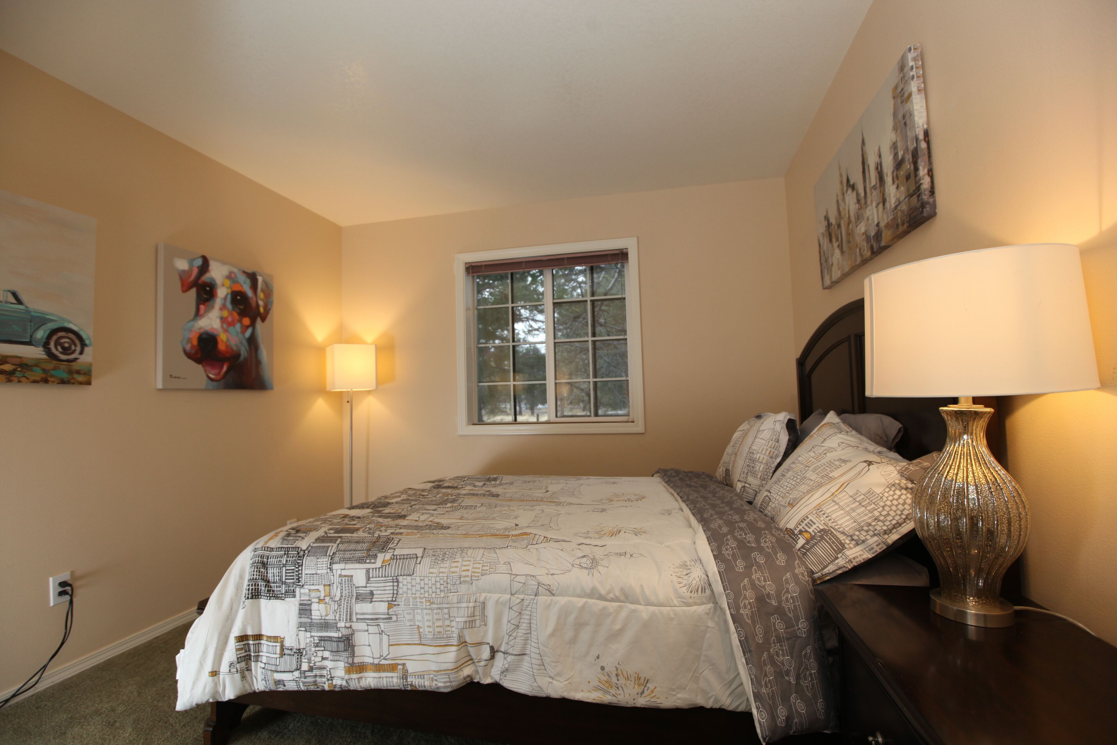 A bedroom with a bed, a window, a lamp, and artwork on the walls.
