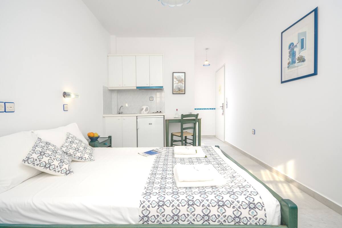 A bedroom with a bed, pillows, a bedside table with a book and a fruit bowl, a small kitchenette, and a chair.