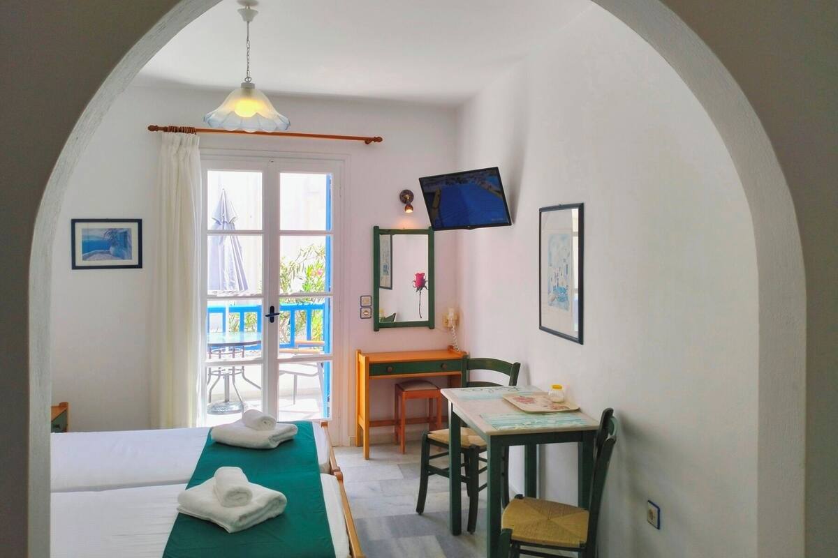 A room with a bed, a desk, a chair, a TV, and a balcony with a blue railing.