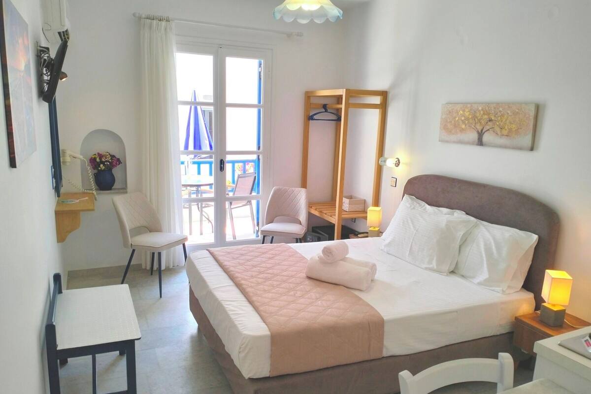 A hotel room with a bed, bedside tables, a desk, a chair, a small table, a mirror, and a balcony with a table and chairs.