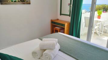 A hotel room with a bed, a desk, a chair, a small table, a green curtain, a potted plant, and a view of the sea.