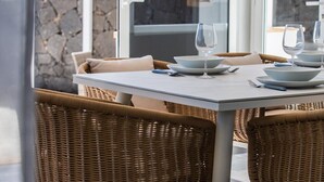 A dining table set with plates, glasses, and cutlery, surrounded by wicker chairs, with a stone wall and a glass door in the background. - Stunning 4 Bedroom Villa on Prestigious Development (Playa Blanca)