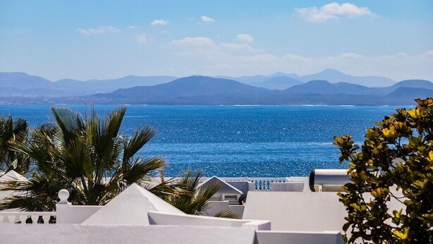 A coastal view with a clear blue sea, white buildings, and palm trees. - Stunning 4 Bedroom Villa on Prestigious Development (Playa Blanca)