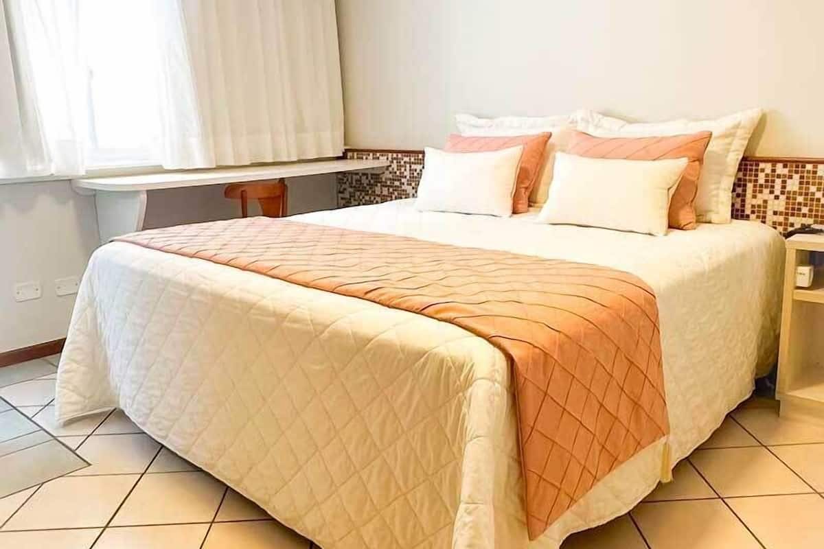 A neatly made bed with a quilted cover, a bedside table with a lamp, and a window with curtains.