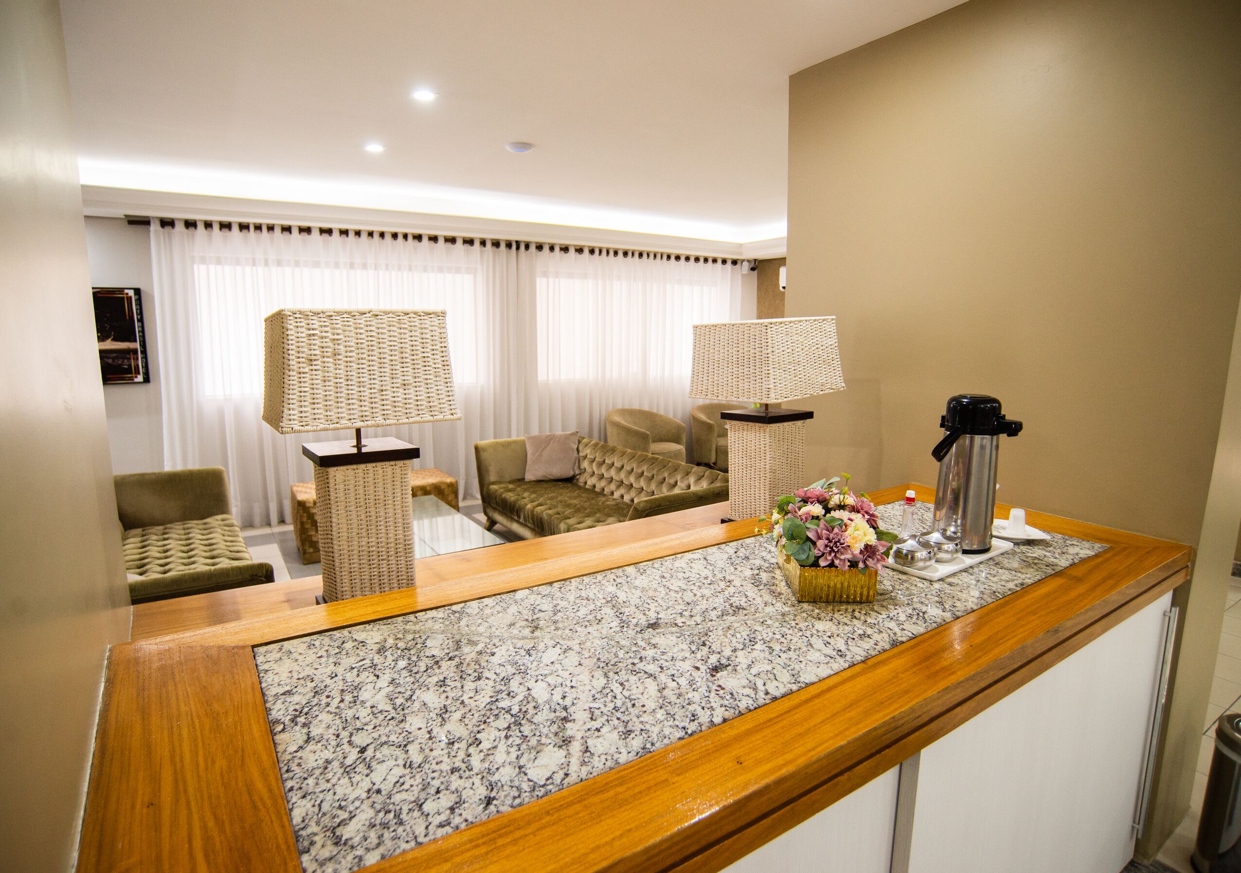 A hotel room with a wooden reception desk, two lamps, a sofa, and a small table with a flower vase.
