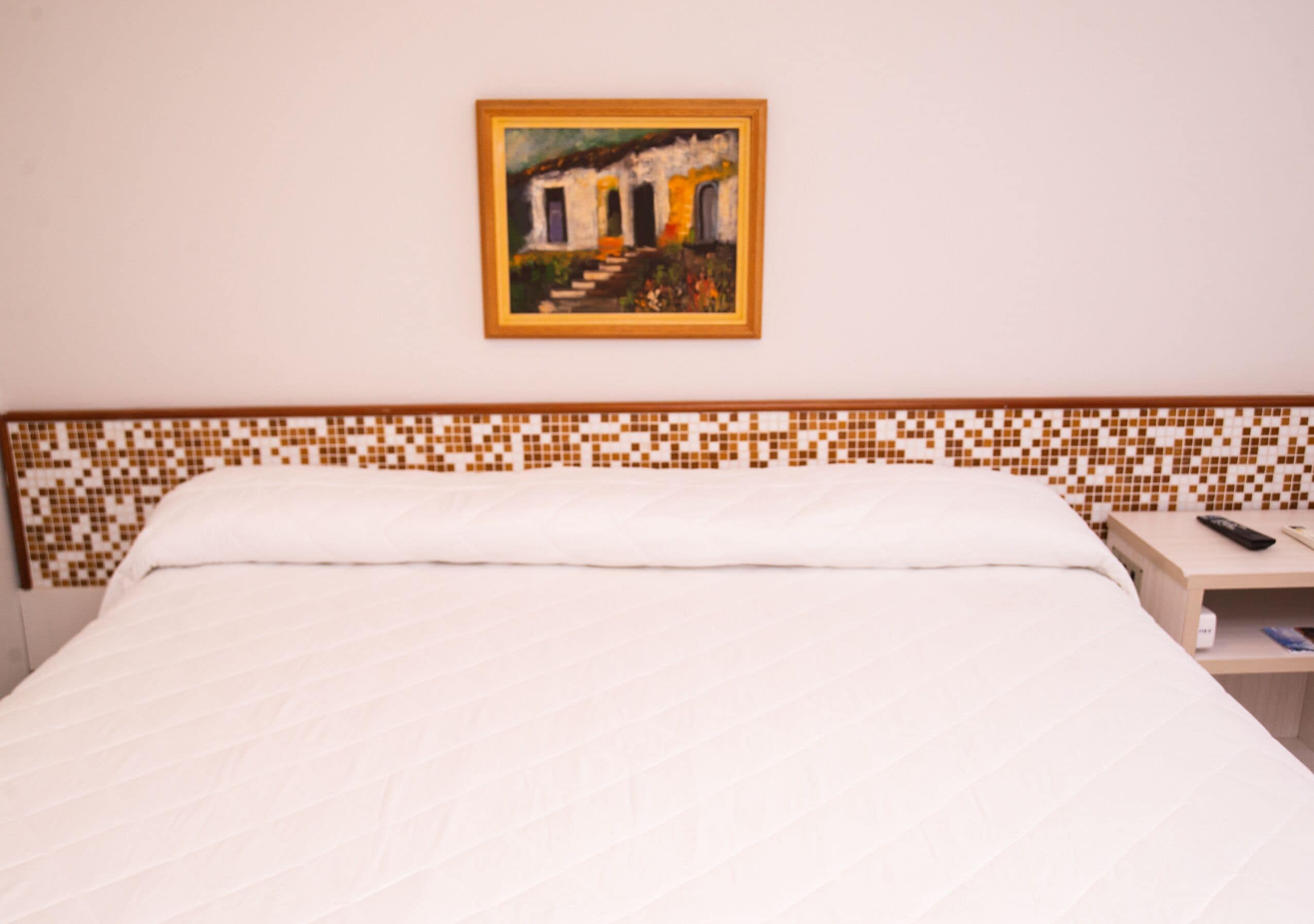 Una habitación de hotel con una cama, una mesita de noche, un control remoto y un cuadro en la pared.