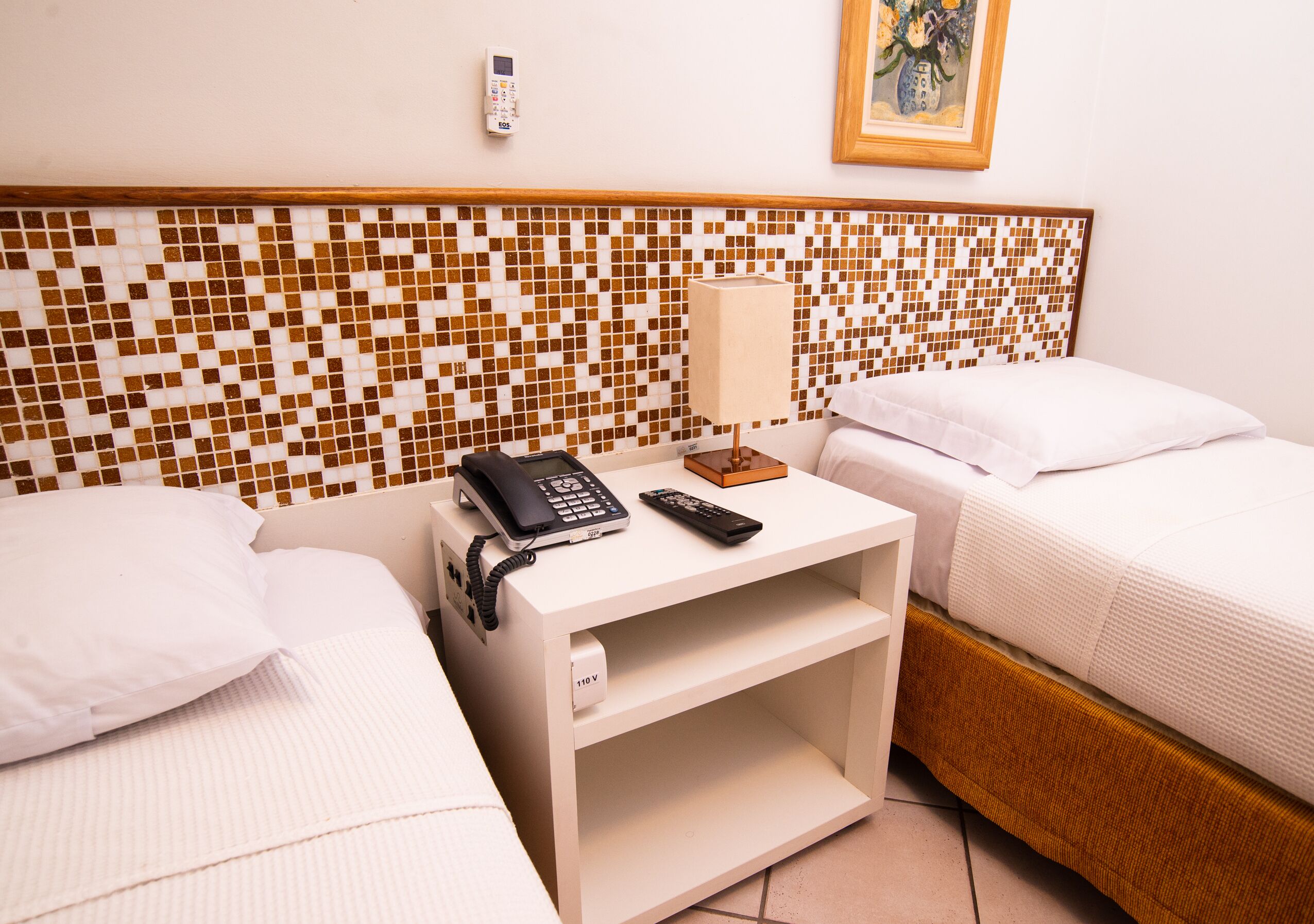 Habitación de hotel con dos camas, una mesita de noche con teléfono y control remoto, un termostato en la pared y un cuadro enmarcado.