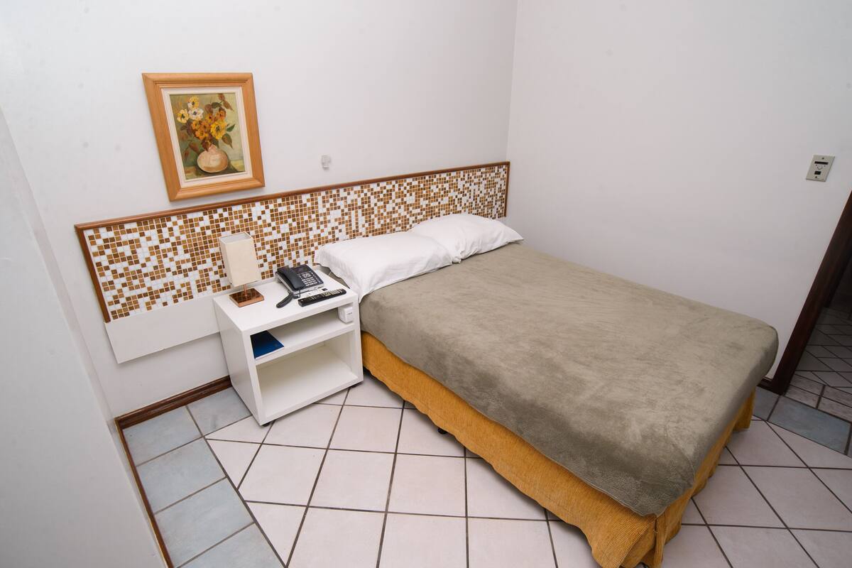 A bedroom with a tiled floor, a bed with a beige blanket, a nightstand with a phone and a remote, and a wall-mounted picture.