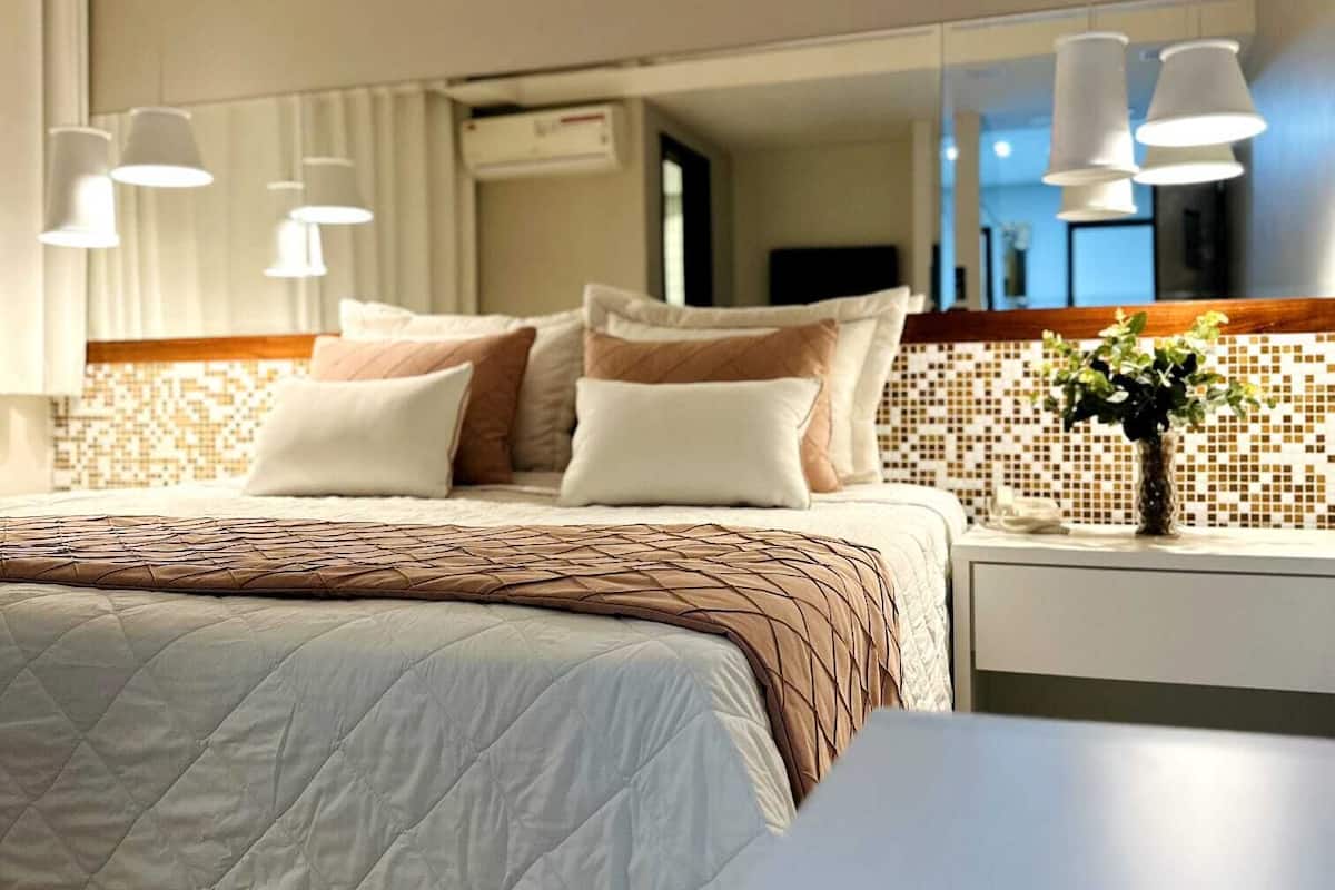 A modern hotel room with a large bed, a bedside table with a vase, and a mirror above the bed.