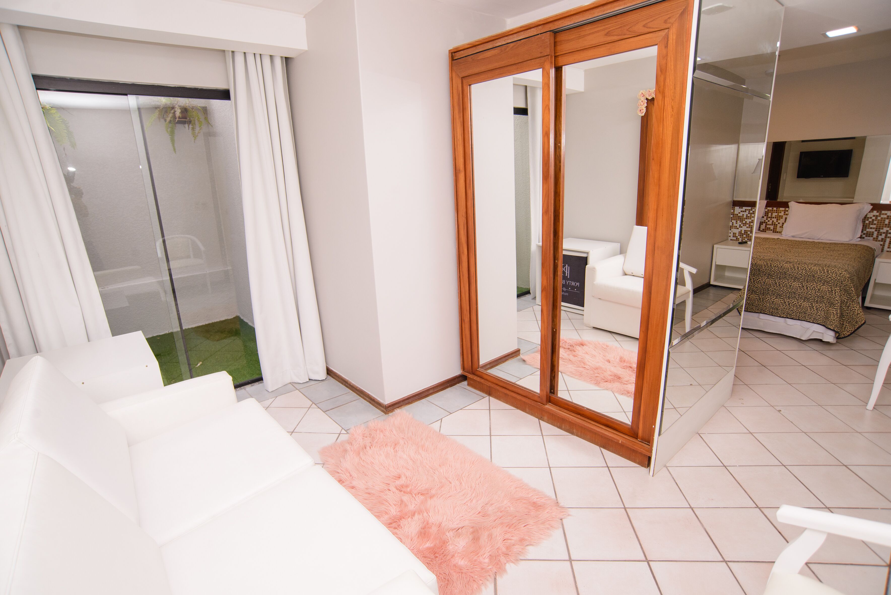 Une chambre d’hôtel moderne avec un canapé blanc, un tapis rose, un grand miroir et un lit recouvert d’une couverture à motifs.