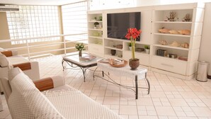 Amplia sala de estar con sofá blanco, una mesita de centro y un televisor en una pared blanca. Se observan estanterías con objetos decorativos y una ventana con vistas.