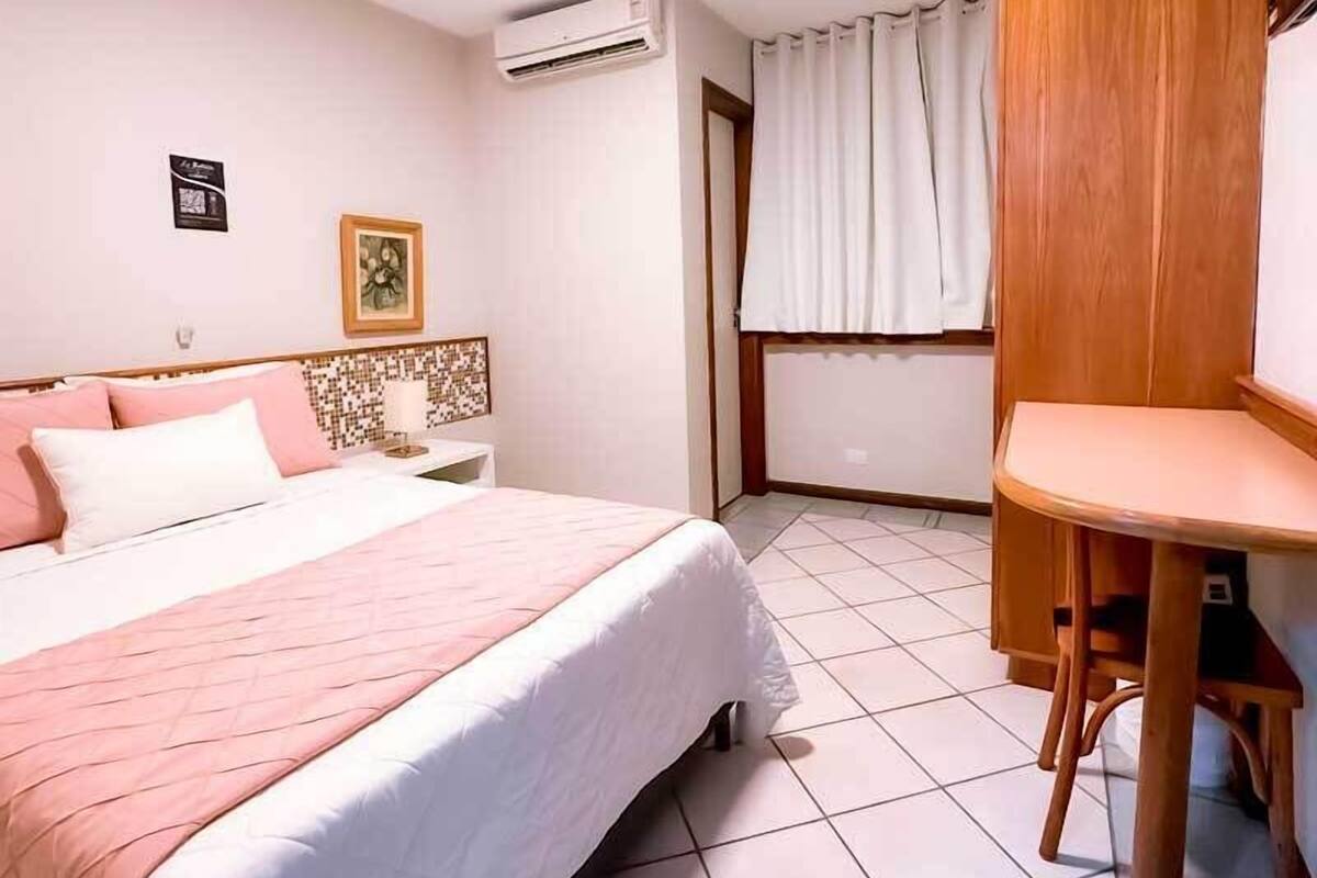 A hotel room with a bed, a bedside table, a chair, an air conditioning unit, and a window with curtains.