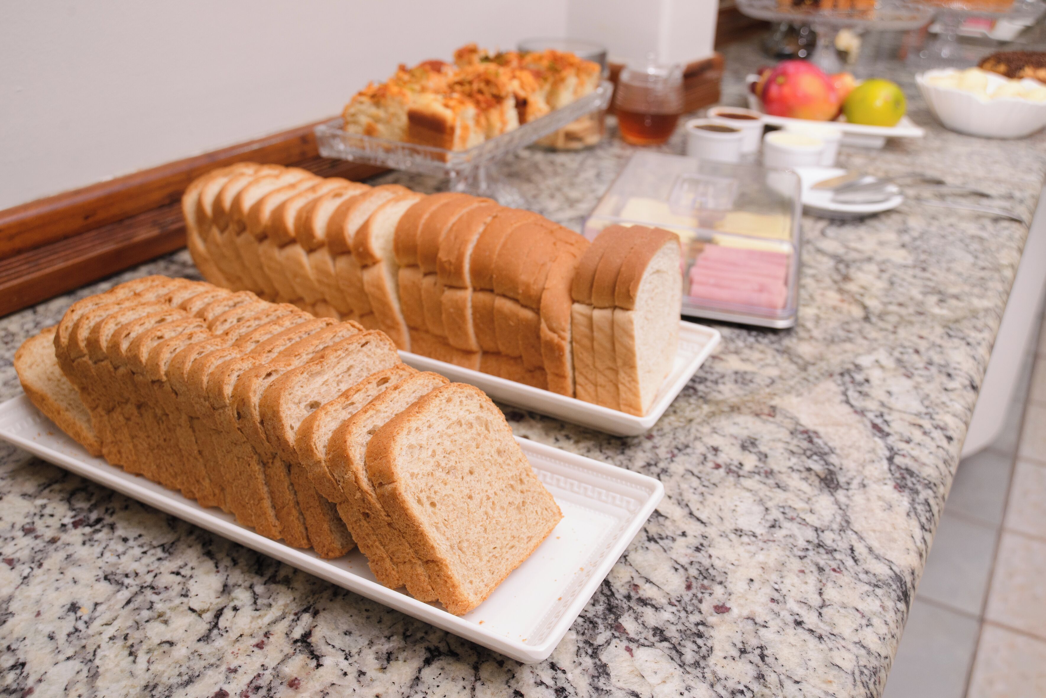 Uma mesa de buffet com pão fatiado, uma bandeja de doces e diversos acompanhamentos.