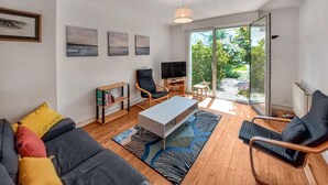 Una sala de estar moderna con un sofá, una mesa de centro, un televisor y una estantería. Hay un ventanal con vista a un jardín.
