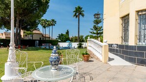 Une terrasse extérieure avec une table en verre, des chaises en métal blanc, un vase décoratif et un palmier en arrière-plan.