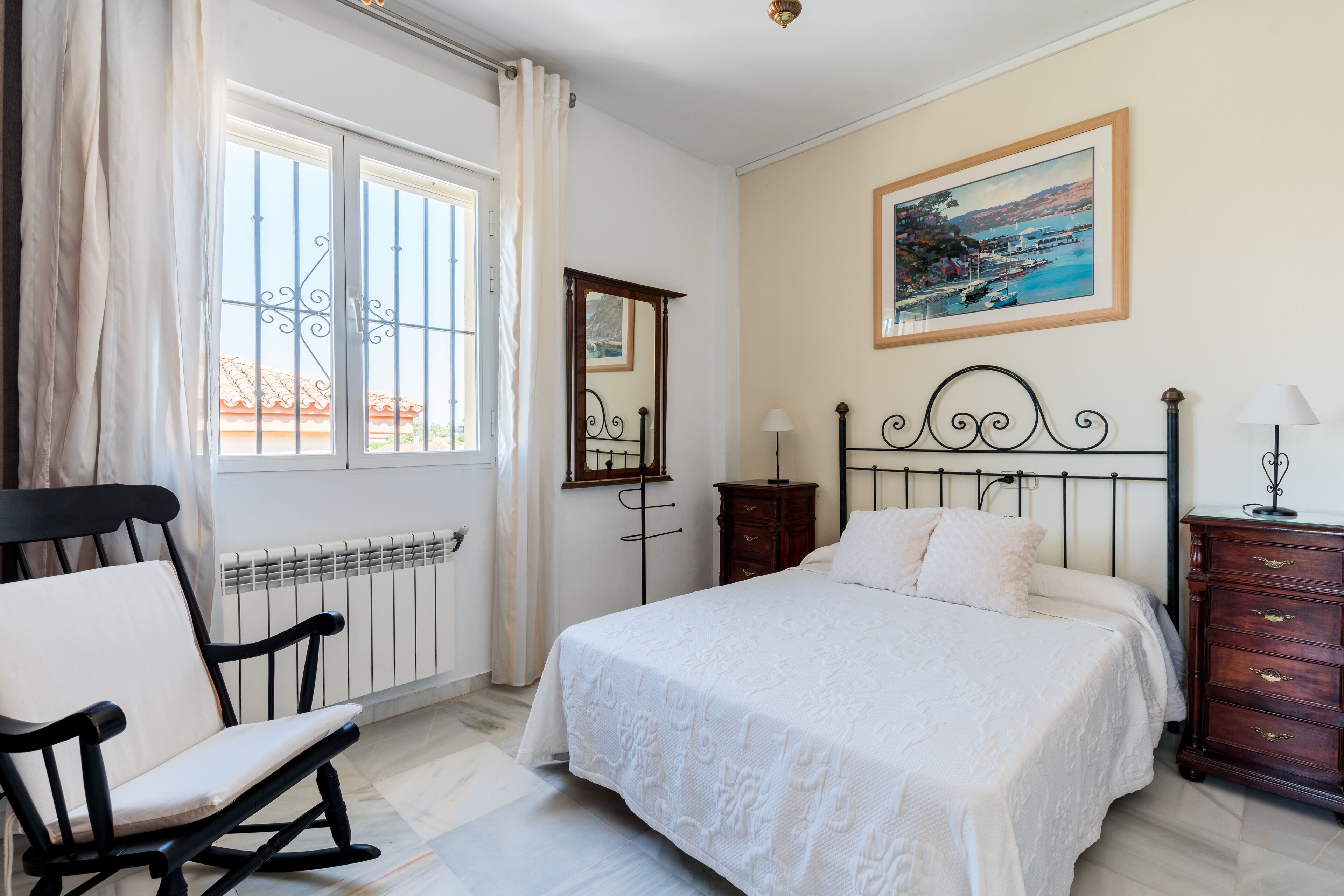 A bedroom with a bed, a chair, a dresser, a mirror, and a window with curtains.