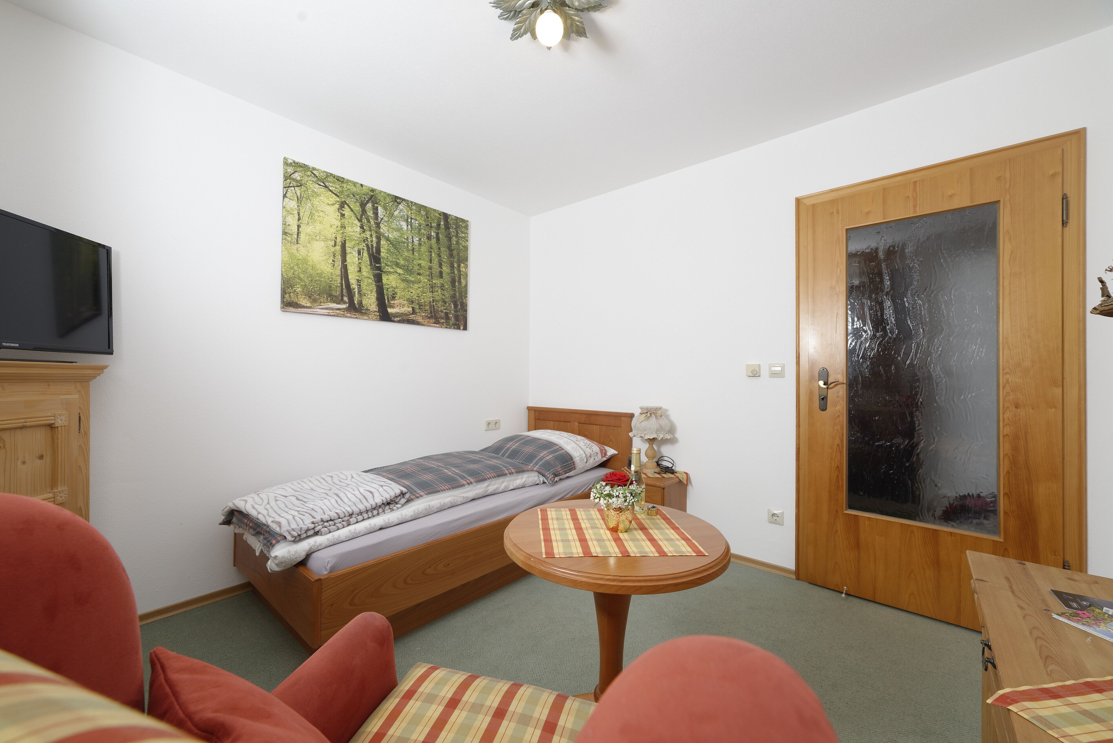 Quarto pequeno e arrumado com uma cama de solteiro, televisão, porta de madeira e uma mesa com abajur.