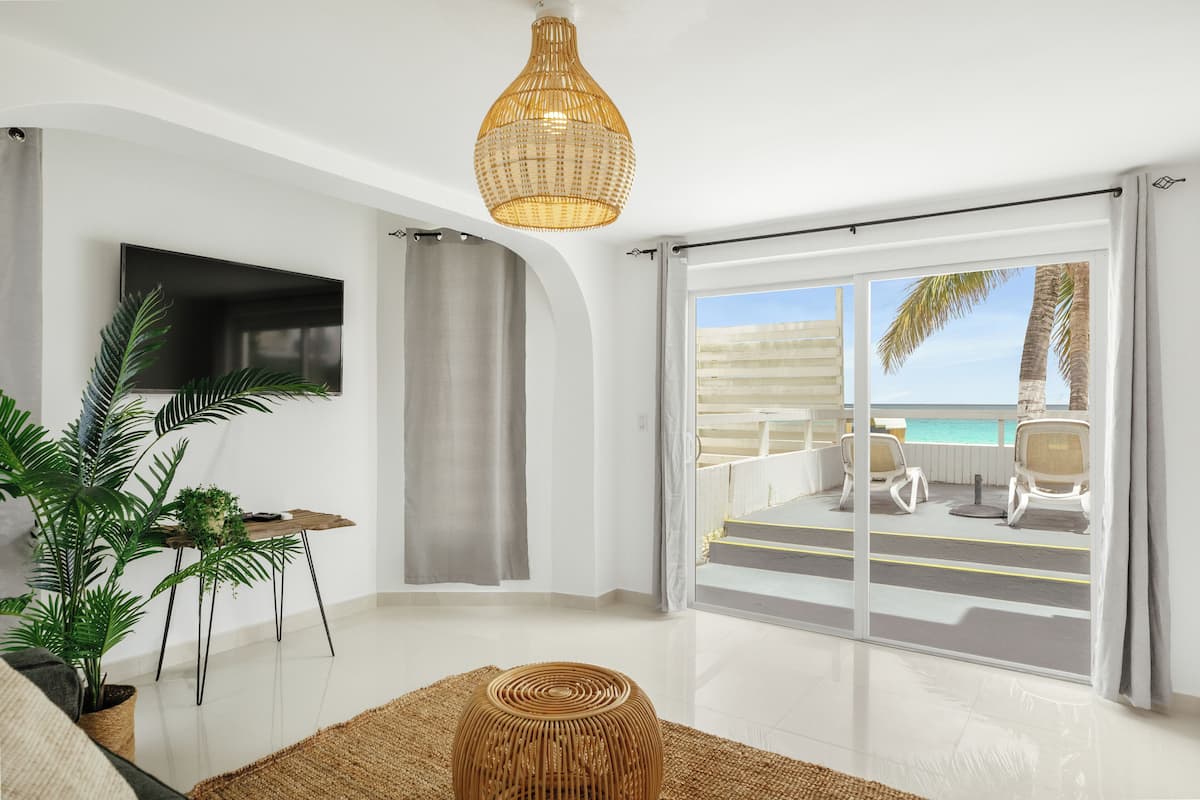 A modern living room with a large TV, a dining table with plants, a wicker ottoman, and a balcony with a view of the ocean.