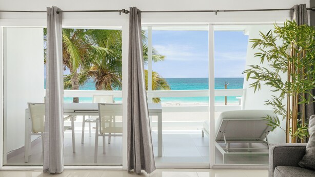 A room with a large sliding glass door leading to a balcony with a view of the ocean and palm trees. - Cocovaa (Simpson Bay)