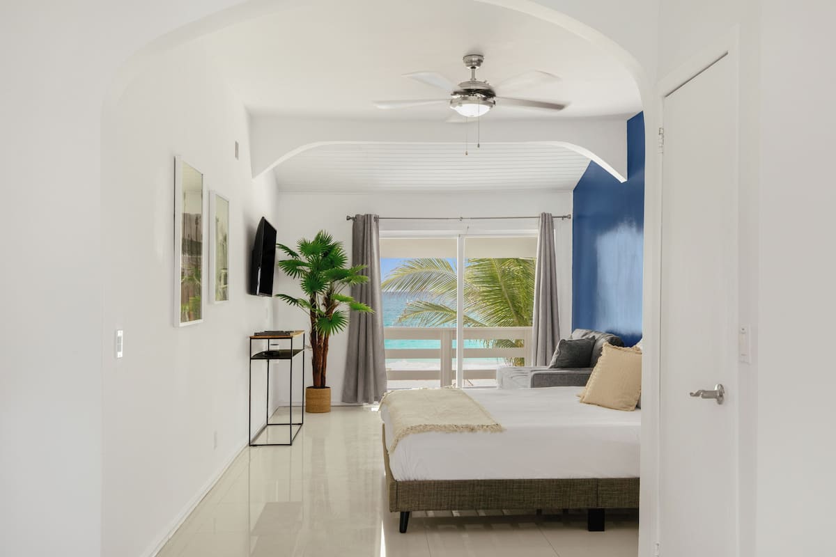 A bedroom with a bed, a ceiling fan, a potted plant, and a view of the ocean through large windows.