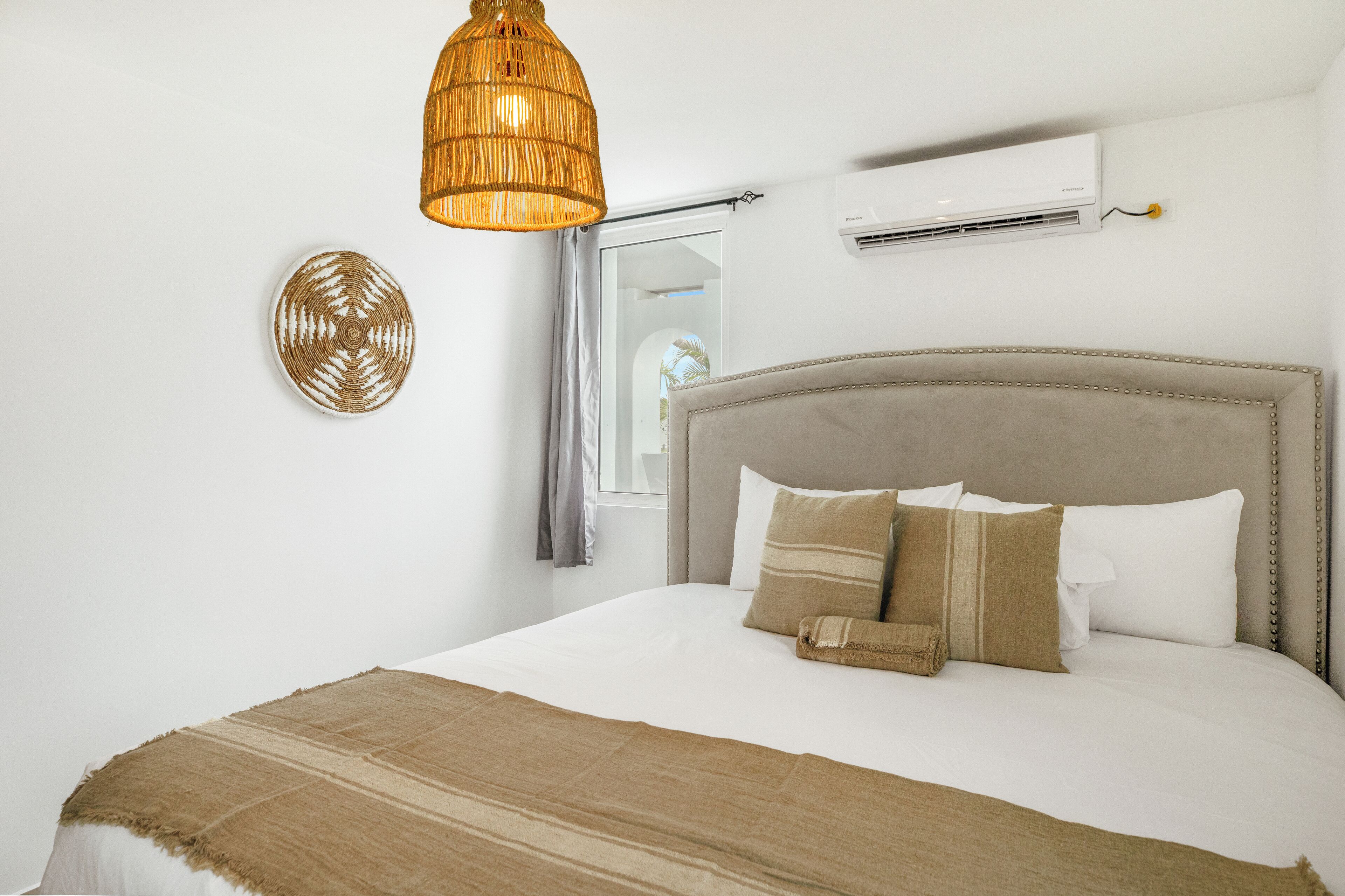A bedroom with a large bed, a hanging light fixture, a wall-mounted air conditioner, and a window with curtains.