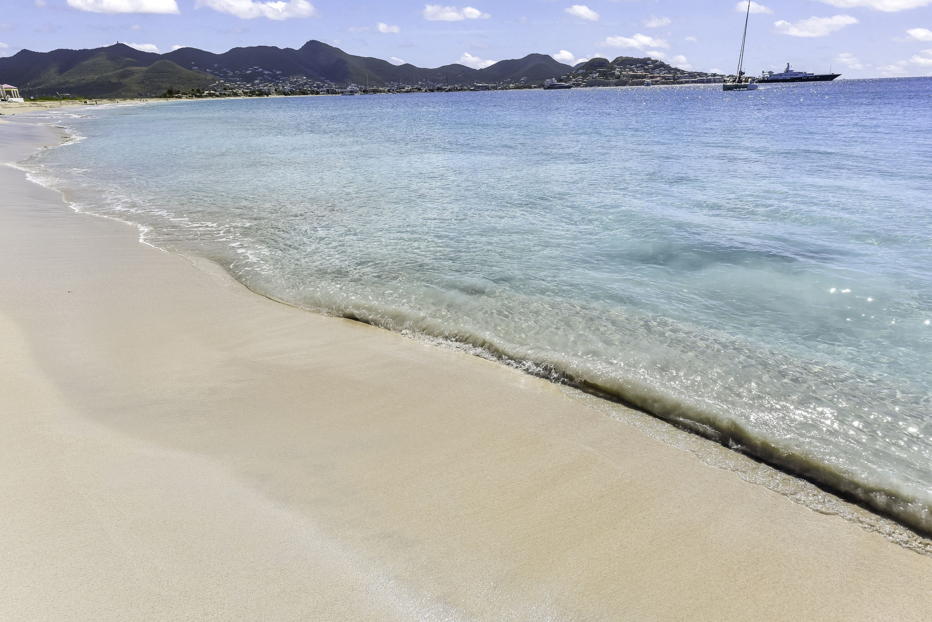 A beach with clear water, a sailboat, and mountains in the background.
